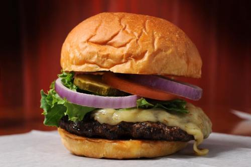 A juicy burger with melted cheese, lettuce, tomato, pickles, and red onion on a toasted bun against a warm background.