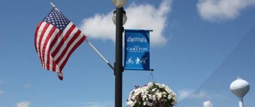 A flag waves beside a blue banner for "The Carlton," with flowers hanging below against a clear sky.