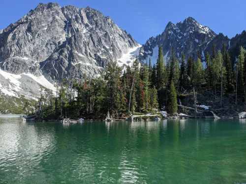 A serene lake surrounded by lush trees and towering mountains under a clear blue sky.