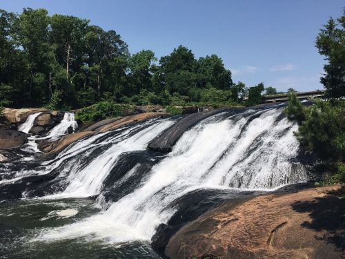 A scenic waterfall cascading over rocky ledges, surrounded by lush green trees under a clear blue sky.