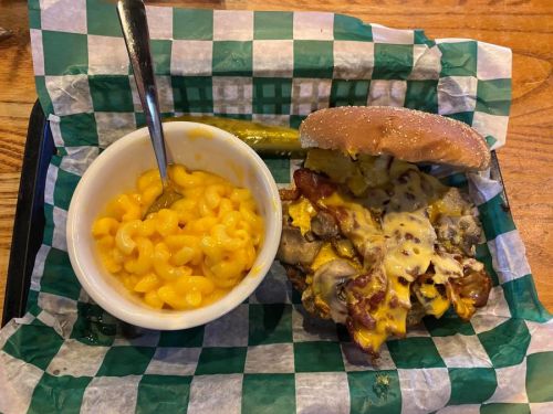 A cheeseburger topped with bacon and mushrooms, served with a side of macaroni and cheese on a checkered tray.