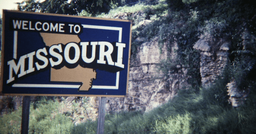 A weathered "Welcome to Missouri" sign stands beside a rocky hillside and overgrown grass.