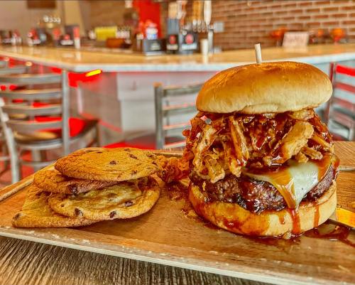 A juicy burger topped with crispy onions and cheese, served with chocolate chip cookies on a wooden platter.