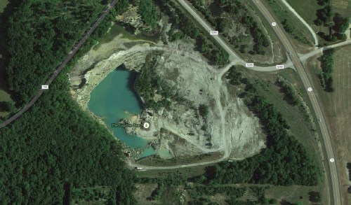 Aerial view of a quarry with a turquoise water-filled pit surrounded by trees and roads.