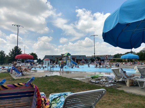 A busy outdoor pool scene with people swimming, lounging on chairs, and colorful umbrellas under a partly cloudy sky.