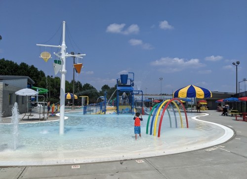 A child stands in a splash pad area with colorful water features and play structures under a clear blue sky.