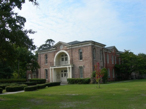 Historic brick building with a central entrance, surrounded by green lawns and trees under a partly cloudy sky.
