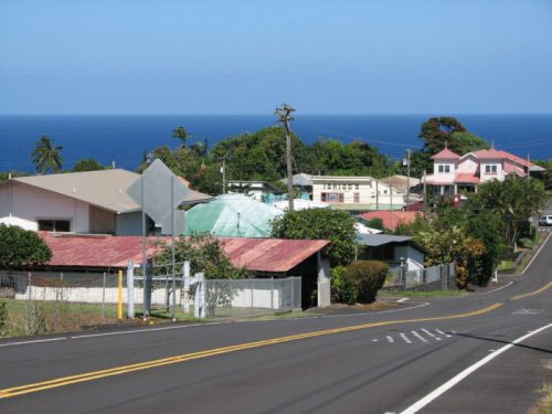 A coastal view of houses along a road, with the ocean and palm trees in the background under a clear blue sky.