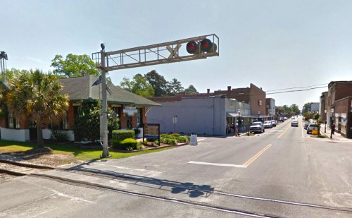 A street view featuring a railroad crossing, shops, and palm trees in a small town setting.