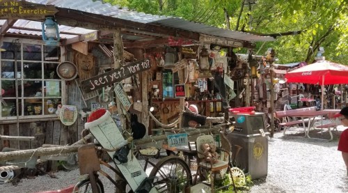 A rustic outdoor scene featuring eclectic shops and decorations, with a bicycle and picnic tables under trees.