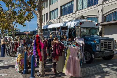 A vintage blue bus parked beside a market, surrounded by people browsing colorful clothing on racks.