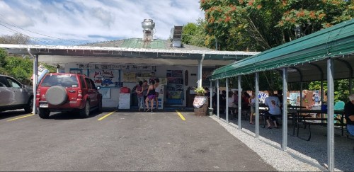 A small roadside shop with outdoor seating, cars parked nearby, and people enjoying refreshments on a sunny day.