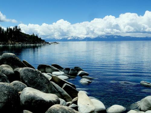 A serene lake scene with smooth rocks in the foreground, calm water, and mountains under a blue sky with fluffy clouds.