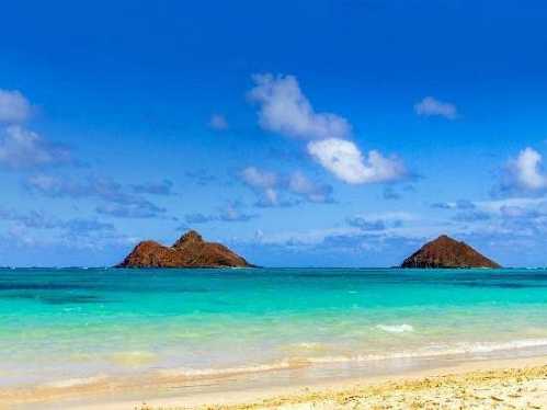 Two small islands rise from turquoise waters under a bright blue sky with fluffy white clouds, viewed from a sandy beach.