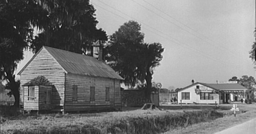 Black and white image of a wooden house and a nearby building, surrounded by trees and a rural road.
