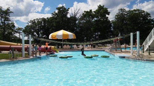 A sunny pool area with water slides, floating mats, and people enjoying the day under a large yellow and white umbrella.