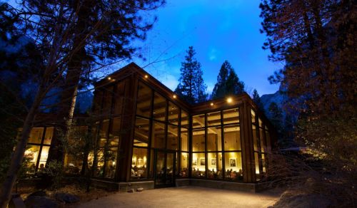 A modern glass-fronted building surrounded by trees, illuminated at dusk in a serene natural setting.