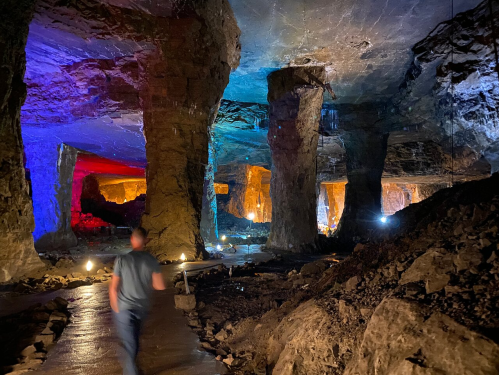 A person walks through a colorful underground cavern with illuminated rock formations and a textured floor.