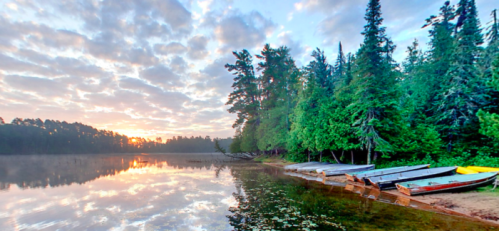 Serene lakeside scene at sunrise, with boats lined up on the shore and trees reflecting in the calm water.