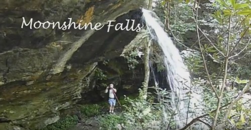 A person stands beneath a waterfall at Moonshine Falls, surrounded by lush greenery and rocky terrain.