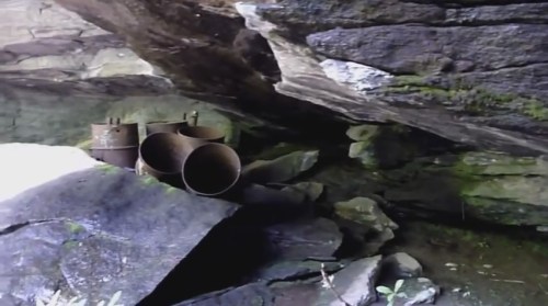 Rusty pipes and barrels sit among rocks under a rocky overhang, with moss and greenery surrounding the area.