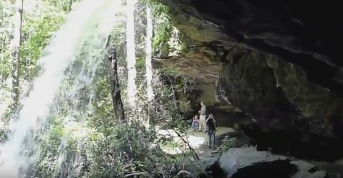 A waterfall cascades over rocks, surrounded by lush greenery, with people exploring nearby.