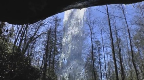 A waterfall cascades down from a rocky overhang, surrounded by trees under a clear blue sky.
