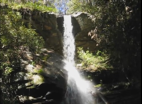 A waterfall cascading down rocky cliffs, surrounded by lush greenery and dappled sunlight.