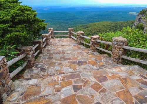 A stone pathway leads to a scenic overlook with lush green hills and a cloudy sky in the background.