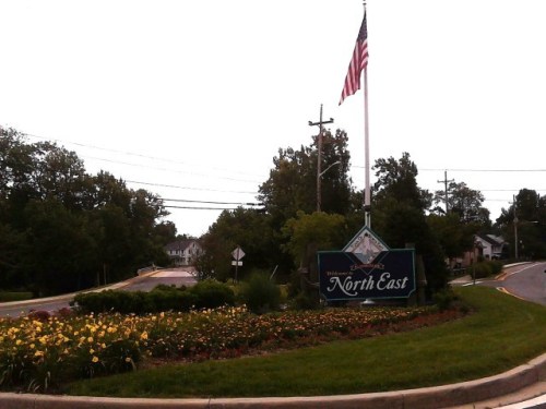 Sign for North East, featuring flowers and an American flag, with a road curving in the background.