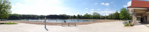 A panoramic view of a sunny park with a sandy beach, a calm lake, and people enjoying the outdoors.