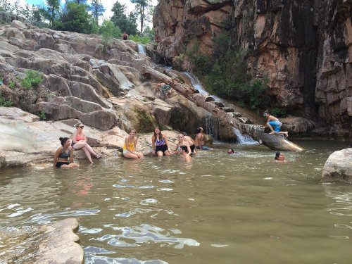 Group of people relaxing in a natural pool surrounded by rocks and a small waterfall in a scenic outdoor setting.