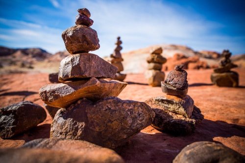 A close-up of stacked stones on a red desert landscape, with more stone formations in the background under a blue sky.