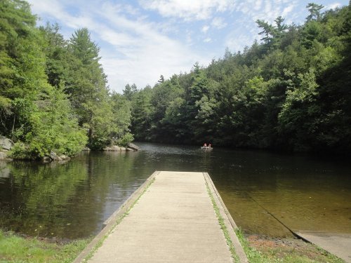 A serene river scene with a concrete dock, surrounded by lush green trees and a small boat in the distance.