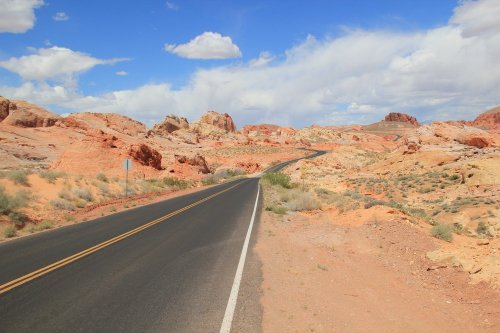 A winding road through vibrant red and orange rock formations under a blue sky with scattered clouds.
