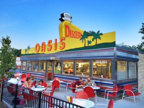 Colorful diner exterior with "Oasis Diner" sign, outdoor seating, and palm trees against a clear blue sky.
