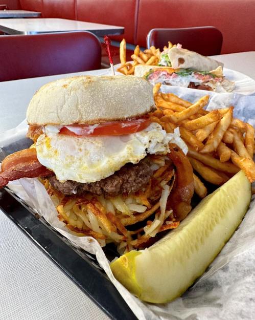A hearty burger with bacon, egg, and hash browns, served with fries and a pickle on a tray in a diner setting.