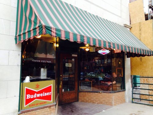 A bar entrance with a striped awning, featuring a Budweiser sign and a cozy interior visible through the glass door.