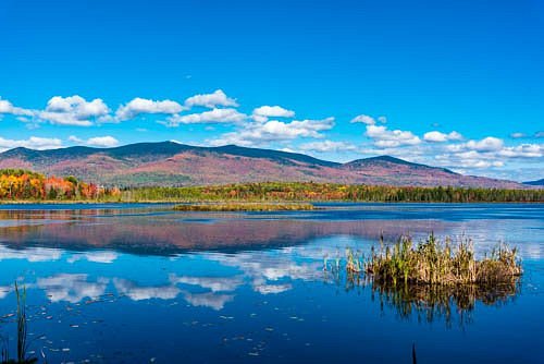 A serene lake reflects blue skies and colorful autumn foliage, surrounded by mountains under fluffy white clouds.