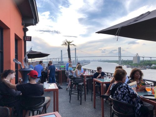 People enjoying drinks on a rooftop terrace with a view of a river and bridge under a cloudy sky.