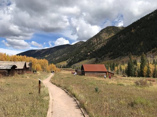 A scenic path through a grassy field, flanked by mountains and rustic cabins, with autumn foliage in the background.