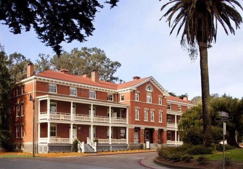 Historic red brick building with multiple balconies, surrounded by trees and palm, under a clear blue sky.