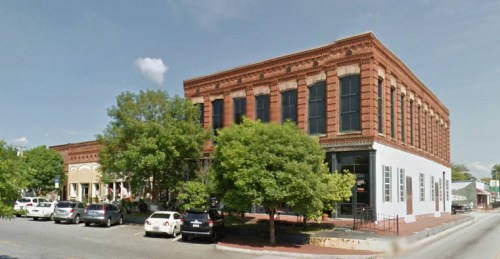 Historic brick building with large windows, surrounded by trees and parked cars on a sunny day.