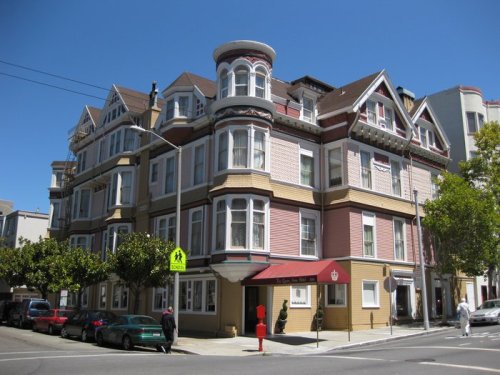 A colorful Victorian-style building with bay windows, located on a sunny street corner, surrounded by trees and parked cars.