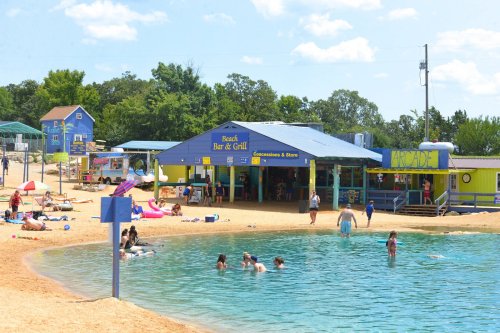 A beach bar and grill by a sandy shore, with people swimming and relaxing under a sunny sky.