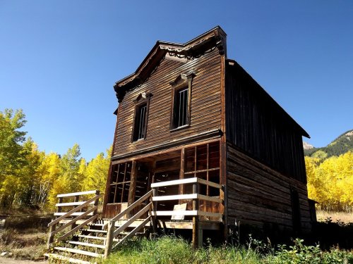 A weathered wooden building with a porch, surrounded by vibrant yellow trees under a clear blue sky.