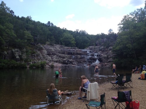 A serene natural scene with people swimming and relaxing by a rocky waterfall and clear water, surrounded by trees.