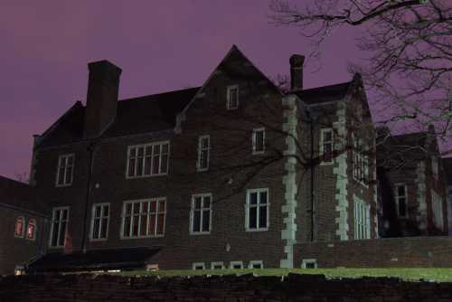 A large, dark brick building with multiple windows, set against a purple sky at dusk. Shadows of trees are visible.