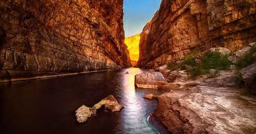 A serene river flows through a narrow canyon, illuminated by warm sunlight reflecting off the rocky cliffs.