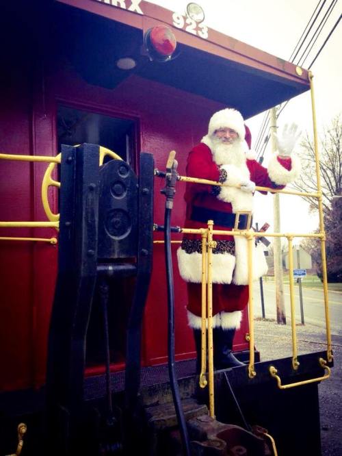 Santa Claus waves from the doorway of a red train caboose, dressed in his traditional red suit and white beard.